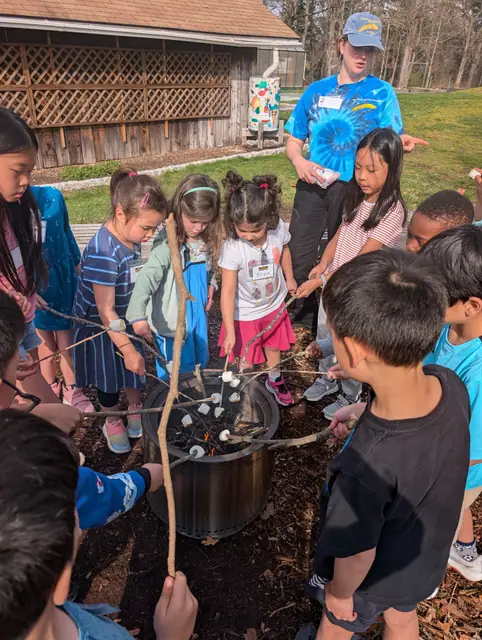 Kids gather around a fire pit with an instructor, each holding a roasting stick.