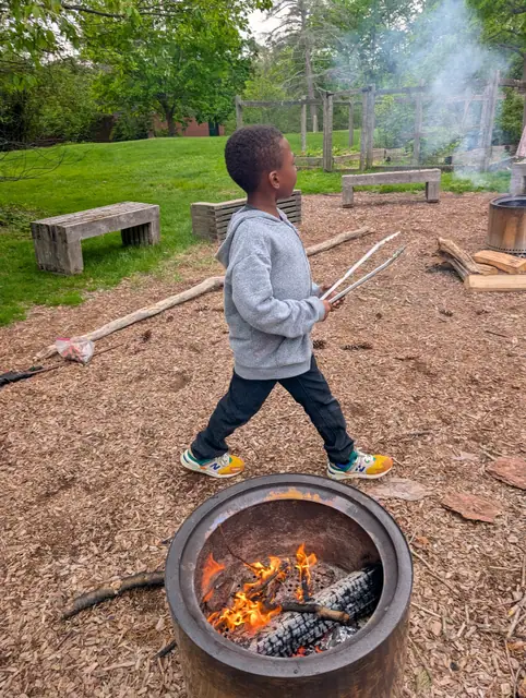 A boy carries two long sticks past a lit fire pit.