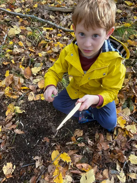 A kid in a yellow raincoat carves a point onto a stick.