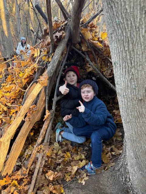 Two kids sit inside a finished lean-to of branches and leaves.