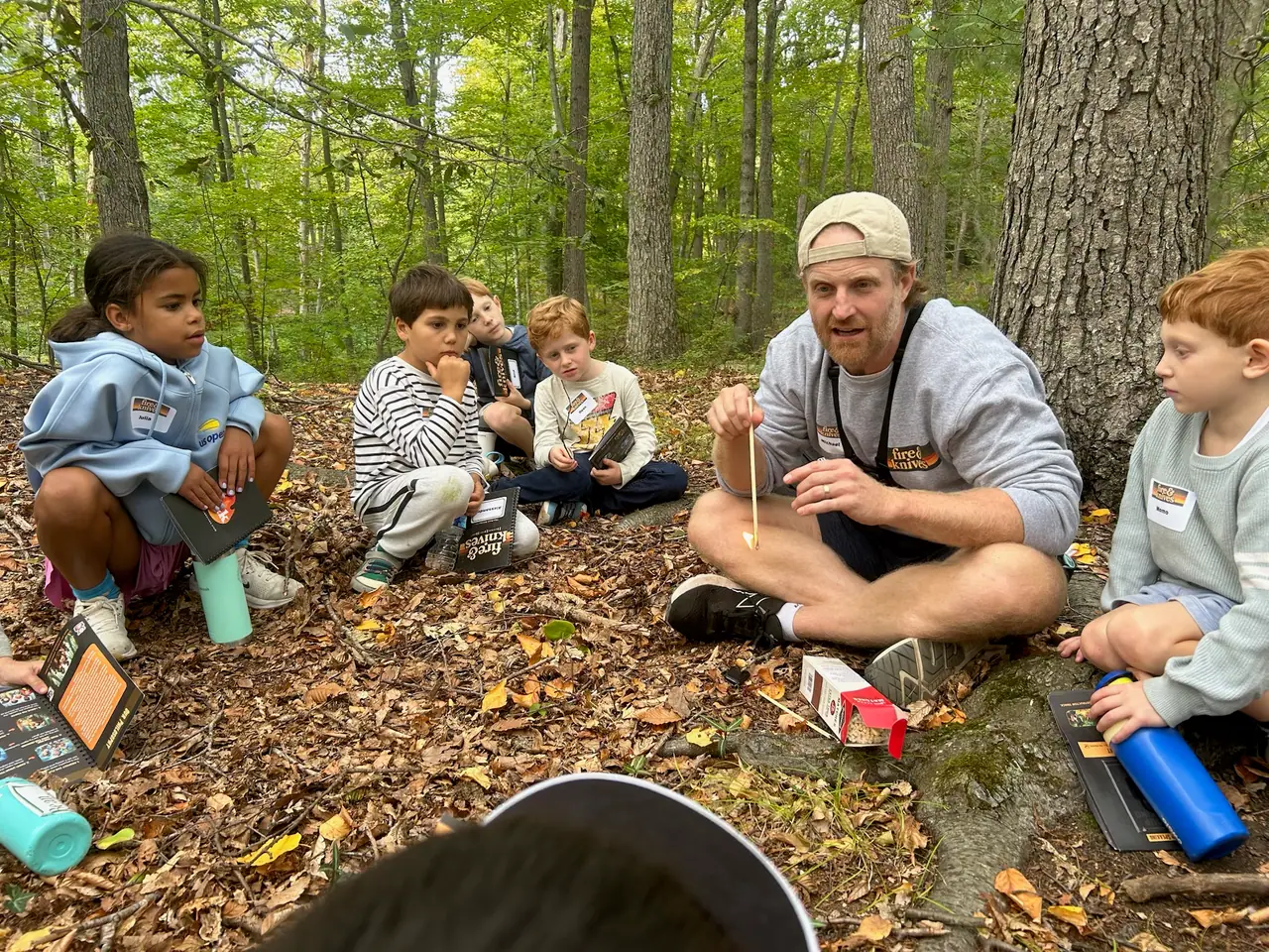 An instructor sits on the ground with a circle of kids in the woods, telling the story of fire before the first strike.